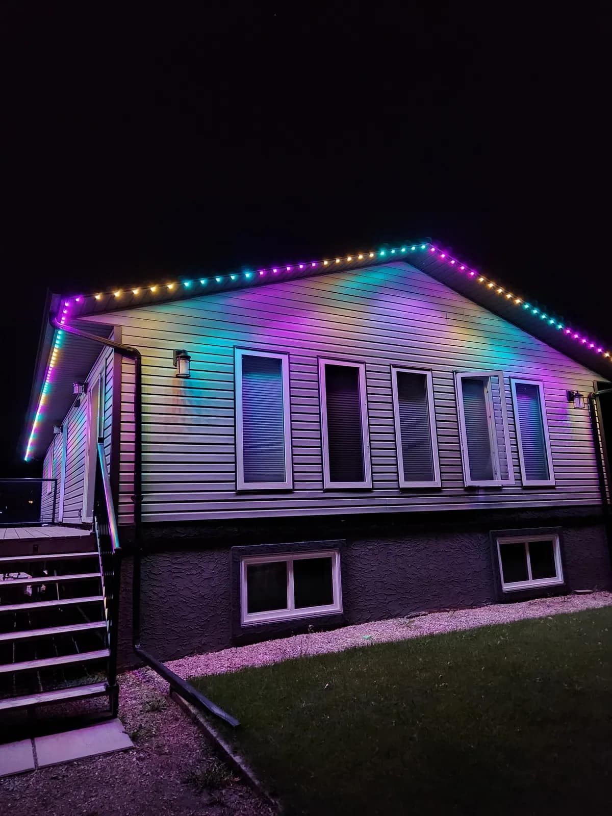 Bungalow with vinyl siding washed in a flowing rainbow gradient from permanent LED roofline lights at night